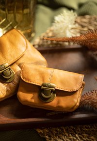 Du keli rudos odinės piniginės su žalvariniais sagtimis, pasižyminčios lygiu paviršiumi ir apvaliais kraštais, padėtos ant medinės lėkštės su dekoratyviniais elementais.