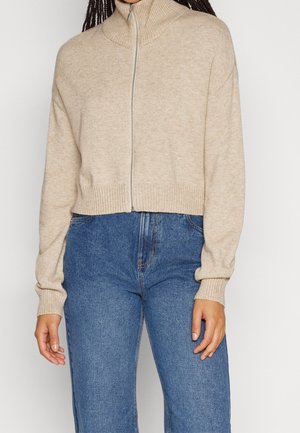 Femme portant un pull en tricot beige zippé avec col et poignets côtelés, associé à un jean bleu taille haute.