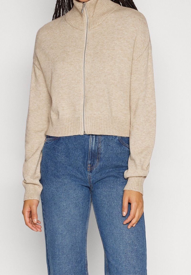 Femme portant un pull en tricot beige zippé avec col et poignets côtelés, associé à un jean bleu taille haute.