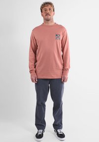 Un homme se tient de face portant une chemise à manches longues corail avec un petit motif floral bleu, un pantalon sombre et des baskets noires avec des lacets blancs.