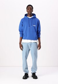 Sudadera azul con cordones en la capucha y bolsillo frontal, superpuesta a una camiseta blanca; combinada con jeans de un azul claro y zapatos negros.