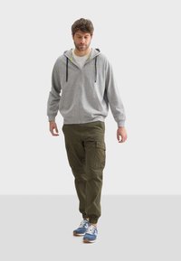 Un sweat à capuche zippé gris avec des cordons de serrage et des accents jaunes, associé à un pantalon cargo olive avec des poches latérales, et des baskets bleues avec des lacets blancs.