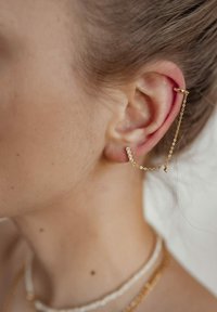 Orecchino d'oro a cuffia con una catena che collega a una barra decorata con cristalli. Design aderente, texture liscia, dettagli minimalisti, fissato dietro l'orecchio.
