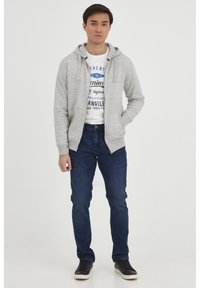 Grå, zip-up hettegenser med to frontlommer, båret over en hvit t-skjorte med grafikk. Mørkeblå jeans og svarte sneakers fullfører antrekket.