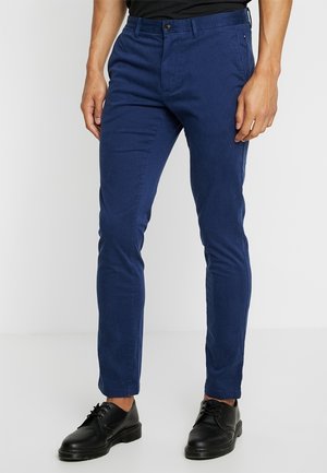 Homme portant un pantalon slim bleu foncé avec des chaussures noires à lacets, debout devant un fond clair uni.