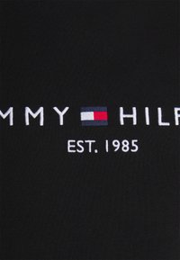 Musta kangas, jossa on brodeerattu teksti "TOMMY HILFIGER" ja vaakasuora väriblokki punaisena, valkoisena ja sinisenä. Teksti sisältää "EST. 1985."