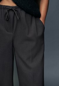 Pantaloni grigio scuro a gamba larga con vita regolabile a coulisse, caratterizzati da una texture liscia e tasche laterali. Il tessuto appare leggero e fluido.