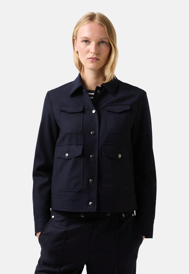 Blonde woman wearing a navy jacket with four flap pockets and silver buttons, paired with matching navy pants, hands in pockets.