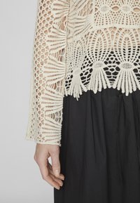 Top in crema all'uncinetto con motivi intricati e tessitura aperta, abbinato a una gonna nera a pieghe. Il dettaglio sulla manica presenta un design testurizzato.