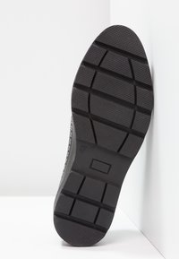 Semelle de chaussure noire avec motif texturé, rainures horizontales et indentations rectangulaires, inclinée contre une surface d’angle blanche.