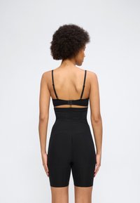 Γυναίκα που φαίνεται από πίσω, φοράει μαύρα σορτς σμίλευσης με ψηλή μέση και μαύρο σουτιέν, με φόντο λευκό και απλό.