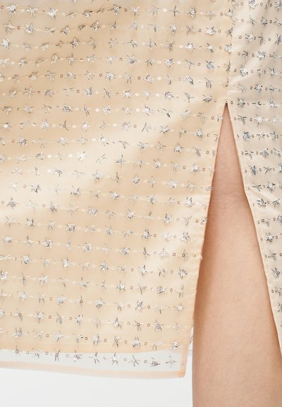 Tissu satin beige orné de motifs floraux argentés brillants, avec une fente latérale pour plus de détails.