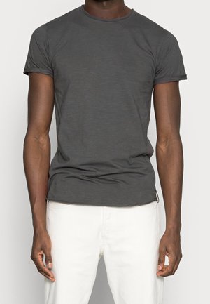 Homme portant un t-shirt ajusté à manches courtes gris foncé et un pantalon blanc, debout devant un fond clair uni.