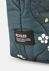Sac en tissu recyclé avec un fond bleu sarcelle foncé orné de motifs floraux blancs et violets, et une étiquette noire indiquant ses matériaux écologiques.