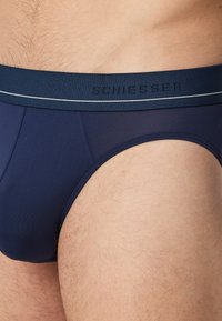 Marineblauwe slip met een zachte elastische tailleband waarop de merknaam "SCHIESSER" staat en een witte accentstreep. Glad oppervlak, aansluitend ontwerp.