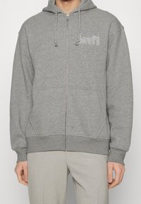 Sudadera gris con cremallera hecha de un tejido suave y texturizado. Presenta capucha con cordón, bolsillos frontales y el logo "Levi's" en blanco.