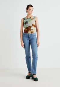 Top brez rokavov v barvah zelenega, rjavega in smetanovega, v kombinaciji z modrimi ravnimi jeans hlačami in zelenimi platformnimi čevlji. Gladke teksture in prilegan dizajn.