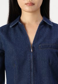 Jeans jumpsuit i mörkblått med dragkedja fram, spetsig krage och bröstficka. Har kontrasterande sömmar och en figurnära silhuett.