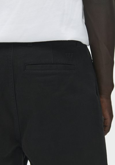 Pantalon noir à texture lisse, avec une poche arrière et un logo "W" en relief discret. Assorti à une chemise blanche portée au-dessus.