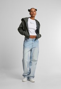 Grønn hettegenser, hvit cropped skjorte med trykk, lyseblå baggy jeans og hvite sneakers. Enkelt design, avslappet passform og myke teksturer.