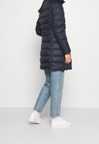 Námořnický modrý péřový kabát s přiléhavým střihem a vertikálním prošíváním; spojený se světlými modrými džíny a bílými teniskami, což vytváří ležérní vzhled.