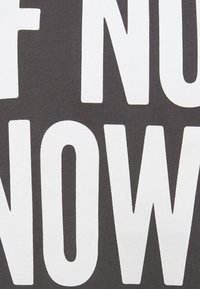 Texto gris con letras en bloque en negrita de color blanco que dicen "SI NO ES AHORA". La tela tiene una superficie suave y ligeramente texturizada.