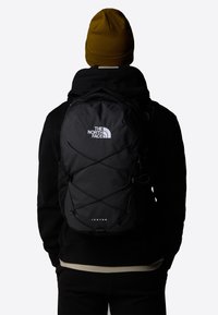 Black backpack with reflective logo and bungee cord design. Features padded straps and ample storage, worn by a person in a dark outfit.