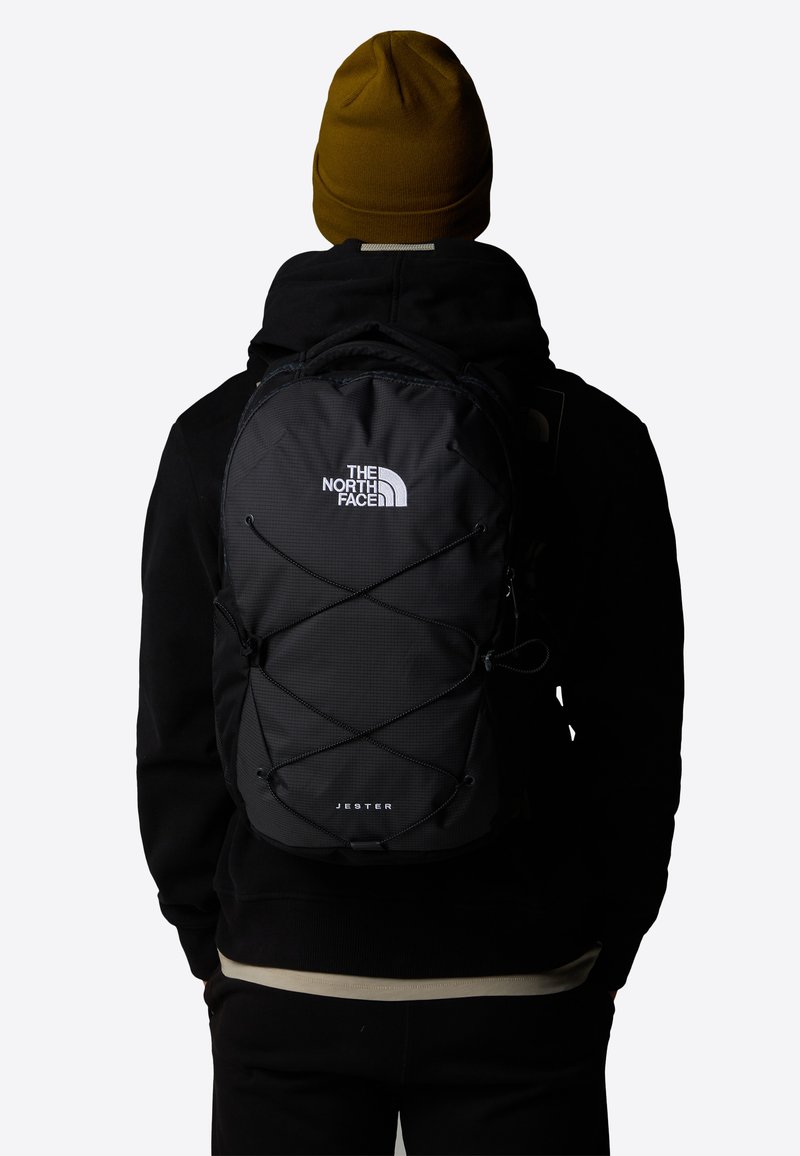 Black backpack with reflective logo and bungee cord design. Features padded straps and ample storage, worn by a person in a dark outfit.