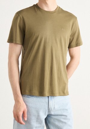 Mann trägt ein olivgrünes T-Shirt mit kurzen Ärmeln und Rundhalsausschnitt sowie hellblaue Jeans, steht mit einer Hand in der Tasche.