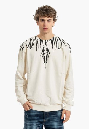 Un jeune homme aux cheveux bouclés porte un sweat-shirt blanc avec un motif abstrait noir autour du col et un jean bleu déchiré.