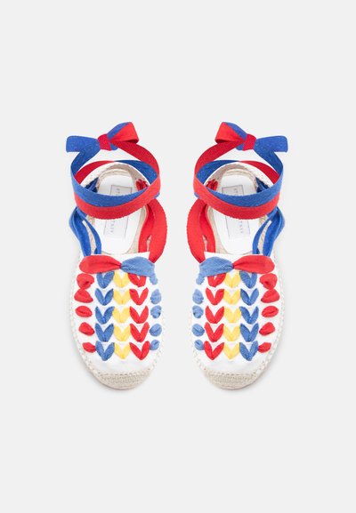 Porų baltų audinių espadrilių sandalų su raudonos, geltonos ir mėlynos spalvos pynimo raštais bei raudonomis ir mėlynomis čiurnos raištelėmis.