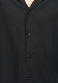 Μαύρο πουκάμισο με κουμπιά, με γιακά με εγκοπή, κλειστό μπροστά με κουμπιά, φορεμένο από άτομο με ορατό δέρμα στον λαιμό.