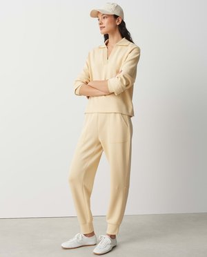 Vrouw staand met gekruiste armen, gekleed in een beige lange mouwen polo, bijpassende joggerbroek, witte sneakers en een beige baseballpet.