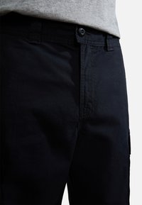 Gros plan d'une personne portant un pantalon noir avec une ceinture boutonnée et des passants pour ceinture, associé à un haut gris clair.
