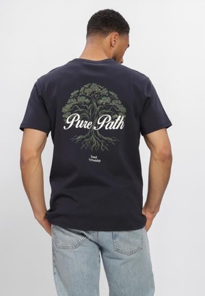 Man wearing black t-shirt with tree graphic and "Pure Path" text, paired with light blue jeans, standing against a plain white background.