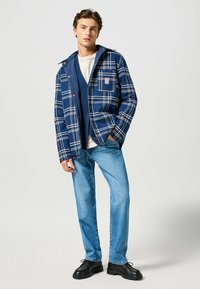 Veste à carreaux marine avec des lignes blanches et bleues, dotée d'une poche poitrine et de boutons, associée à un jean bleu clair et des chaussures noires.