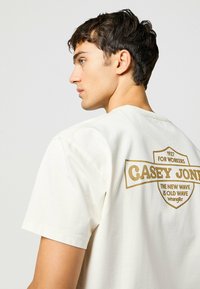 Camiseta de algodón blanca con mangas cortas. Presenta un gran logo impreso en dorado en la parte posterior: "CASEY JONES FOR WORKERS." Diseño sencillo en general.