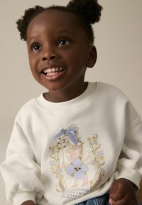Weißer Sweater mit geripptem Ausschnitt und Bündchen, verziert mit floralen Stickereien in Pastellfarben und einem lavendelfarbenen Schleifenakzent auf der Vorderseite.