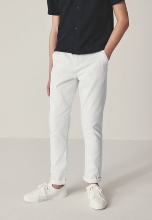Personne portant un pantalon blanc retroussé, des baskets blanches et une chemise noire à manches courtes boutonnée, les mains dans les poches.