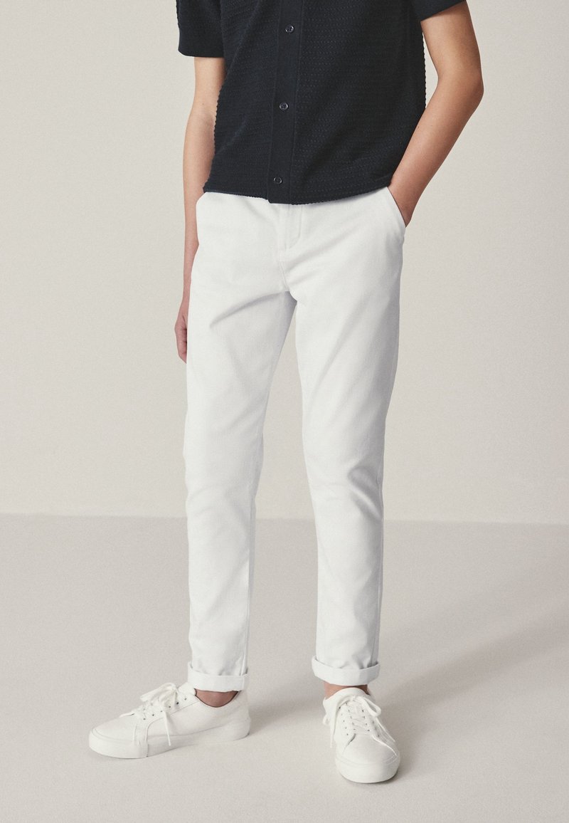 Personne portant un pantalon blanc retroussé, des baskets blanches et une chemise noire à manches courtes boutonnée, les mains dans les poches.