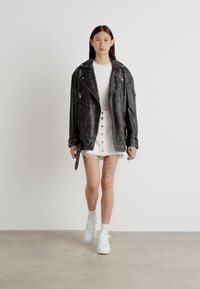 Schwarze Lederbikerjacke mit silbernen Beschlägen, getragen über einem weißen T-Shirt und einem ausgefransten weißen Denim-Rock, kombiniert mit weißen Sneakers.