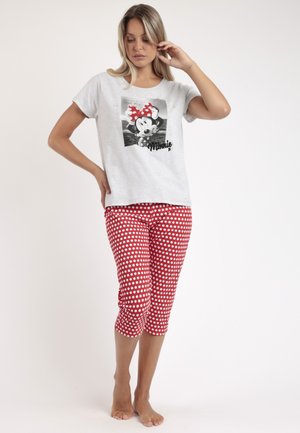 Mujer con camiseta gris de Minnie Mouse y pantalones capri rojos con lunares blancos, de pie descalza frente a un fondo liso.