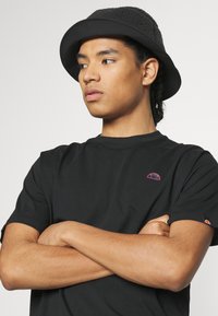 Black short-sleeve t-shirt features a small pink logo on the chest. The model also wears a black textured bucket hat. Arms crossed.