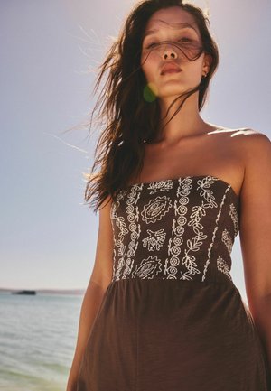 Jauna moteris su palaidais tamsiais plaukais, dėvinti be petnešėlių raštuotą rudą suknelę, stovinti prie vandens po skaidriu dangumi su saulės šviesa.