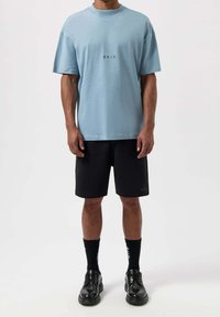 Hellblaues T-Shirt mit kurzen Ärmeln und Rundhalsausschnitt, mit einem kleinen Logo auf der Brust, kombiniert mit schwarzen Shorts und schwarzen Schuhen.