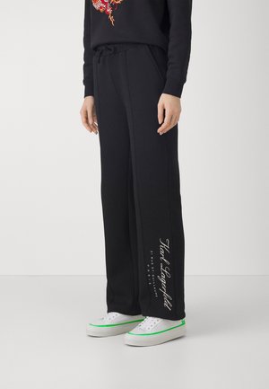 Osoba nosząca czarne spodnie o szerokich nogawach z białym logo Karl Lagerfeld na lewej nodze oraz białe trampki z zielonym obszyciem.