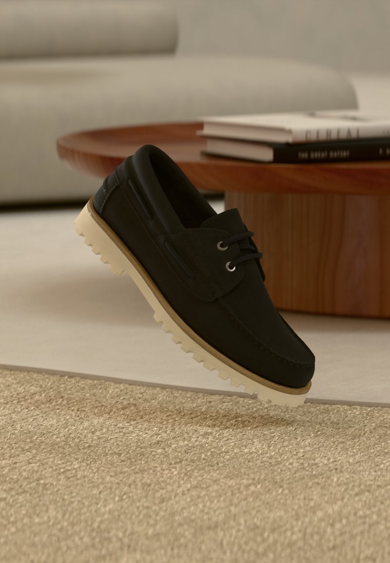 Svart mokkasinstil sko med beige robust såle svever over beige vevd teppe, rundt bord i tre med stablede bøker i bakgrunnen.