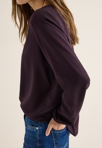 Fioletowa bluzka z długim rękawem z lekkiego materiału, o luźnym kroju, okrągłym dekolcie i elastycznych mankietach, w połączeniu z niebieskimi dżinsami.