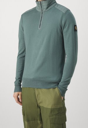 Mann trägt einen türkisfarbenen Pullover mit Viertelreißverschluss, gerippten Bündchen und Kragen, kombiniert mit zweifarbig grüner Cargohose, steht mit den Händen in den Taschen.