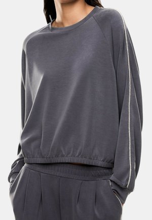 Donna che indossa un top grigio scuro a maniche lunghe e pantaloni abbinati con vita elastica e una striscia argentata lungo le maniche. Mani nelle tasche.